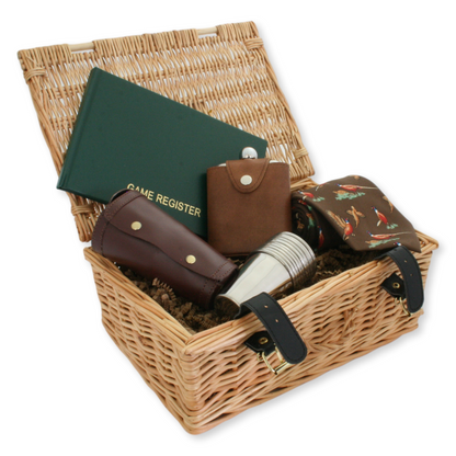 A wicker basket gift set containing a stainless steel hip flask in a leather case, numbered stirrup cups, a brown pheasant tie, and a game register.