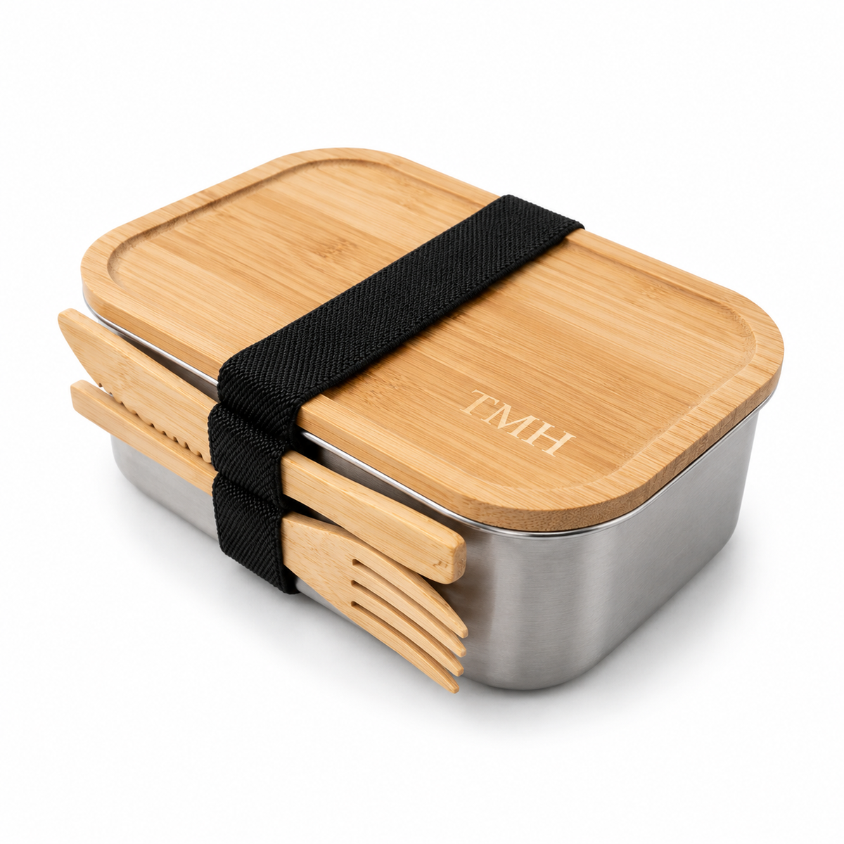 A stainless steel bento lunch box with a bamboo lid, secured by a black elastic band holding matching bamboo utensils. The lid is personalized with the initials 'TMH' in gold, set against a clean white background.