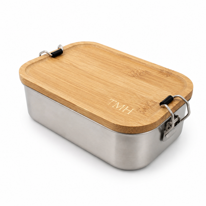 A rectangular stainless steel lunch box with a light natural bamboo lid featuring the engraved initials 'TMH'. The box has black plastic and metal latch closures on the sides, shown on a clean white background.