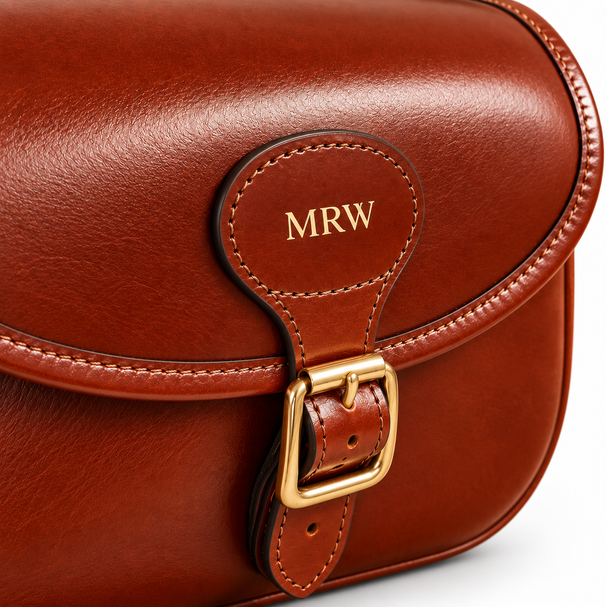 Close-up of a brown leather cartridge bag with a gold-tone buckle and a personalized flap tab stamped with the initials 'MRW'.