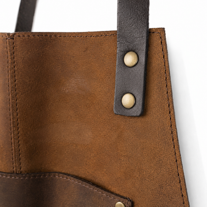 Close-up of a brown leather gunsmith's apron showing stitched panels, a dark brown strap, and brass-tone rivets on a white background.