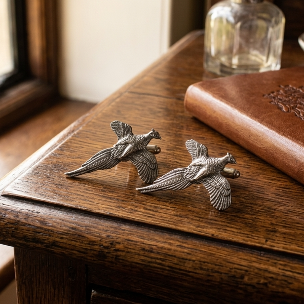 A pair of silver-toned metal cufflinks shaped like pheasants, displayed on a dark wooden table next to a brown leather-bound book and a glass decanter.