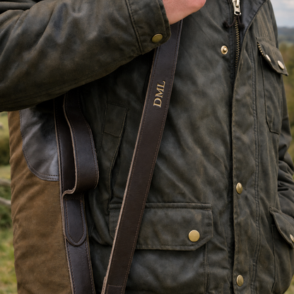 Person wearing a dark green jacket with a brown strap of a gun slip over his shoulder, outdoors.