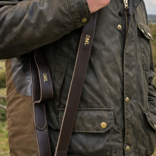 Person wearing a dark green jacket with a brown strap of a gun slip over his shoulder, outdoors.