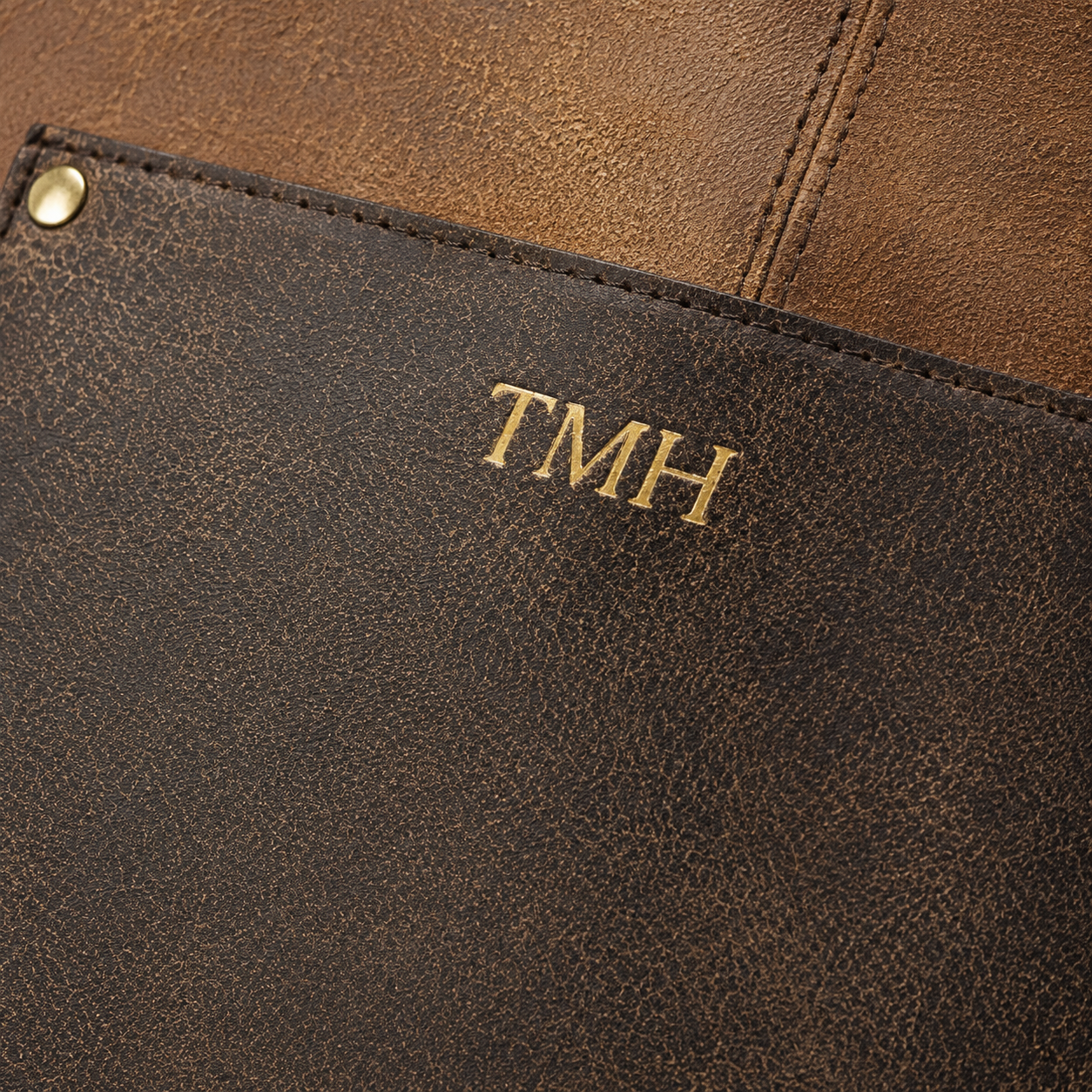 Close-up of a dark brown textured leather gunsmith's apron with gold-tone metal snap and gold-embossed initials 'TMH' on a brown leather background.