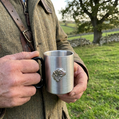 Person holding a stainless steel mug with a gundog emblem in a countryside setting