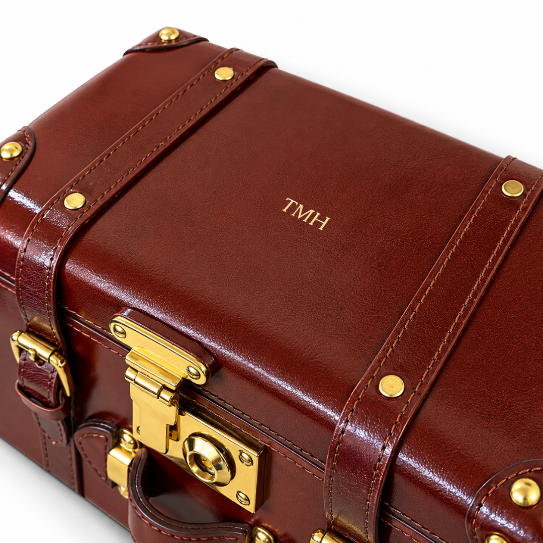 A close-up of a brown leather suitcase or travel case with gold-tone hardware, brass rivets, and a personalized monogram 'TMH' on the front, set against a clean white background.