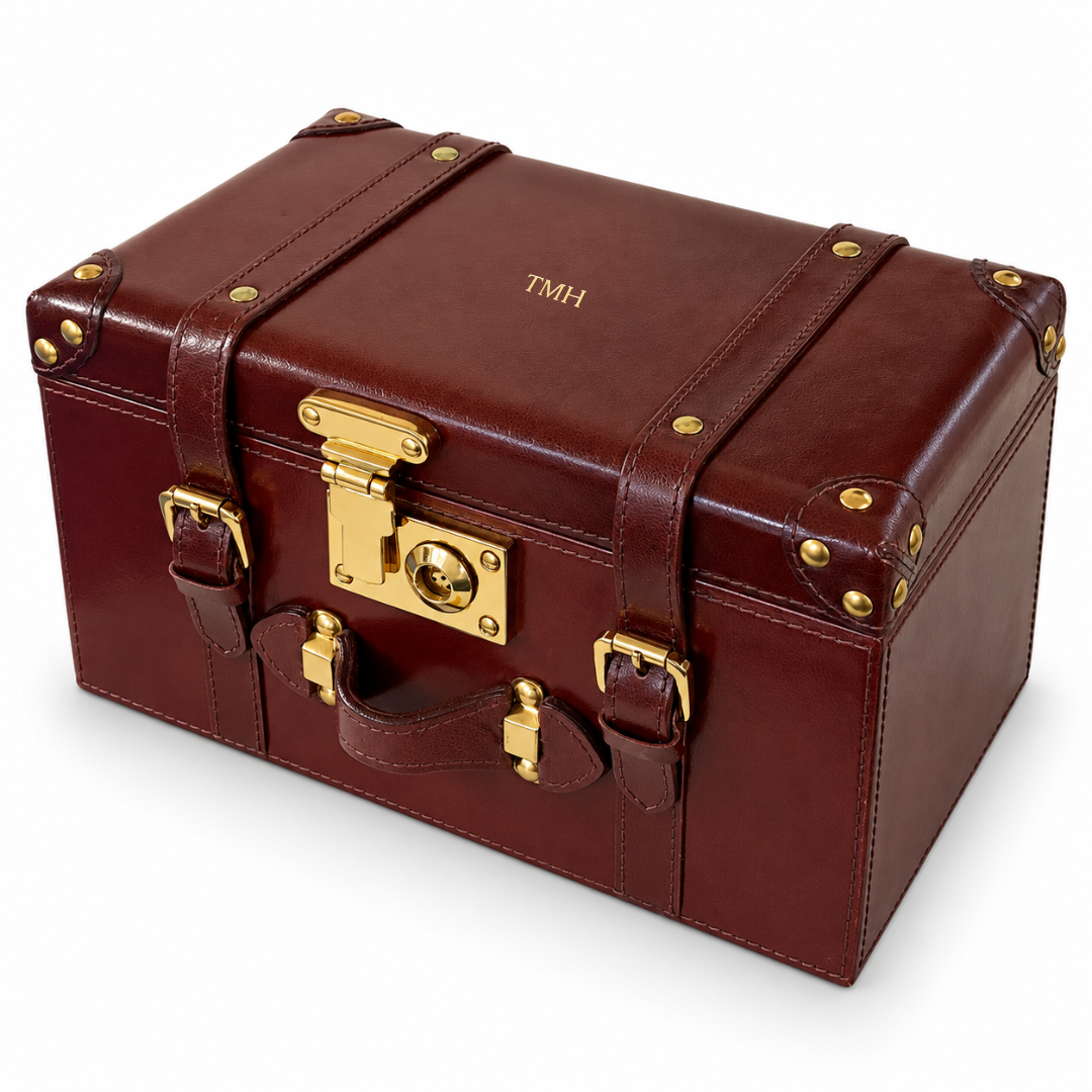 A vintage-style brown leather storage trunk with gold-tone metal hardware, including a latch, straps, studs, and corner protectors, shown on a white background.