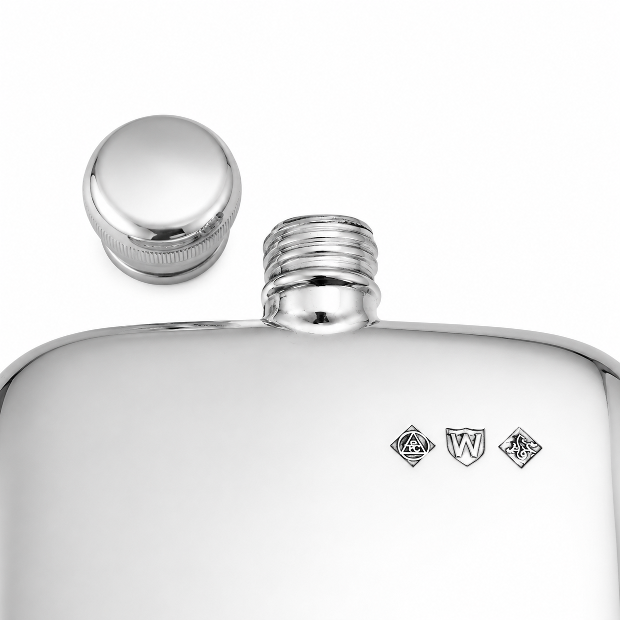 A close-up of a polished silver metal flask with its cap removed and placed beside the threaded neck. Small engraved heraldic marks are visible on the front.
