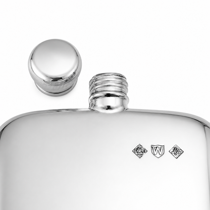 A close-up of a polished silver metal flask with its cap removed and placed beside the threaded neck. Small engraved heraldic marks are visible on the front.