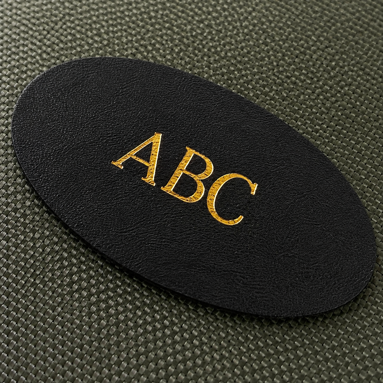 A close-up of a black oval patch with the letters 'ABC' in metallic gold foil, placed on a textured dark olive green fabric rifle cleaning kit