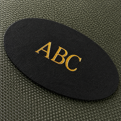 A close-up of a black oval patch with the letters 'ABC' in metallic gold foil, placed on a textured dark olive green fabric rifle cleaning kit
