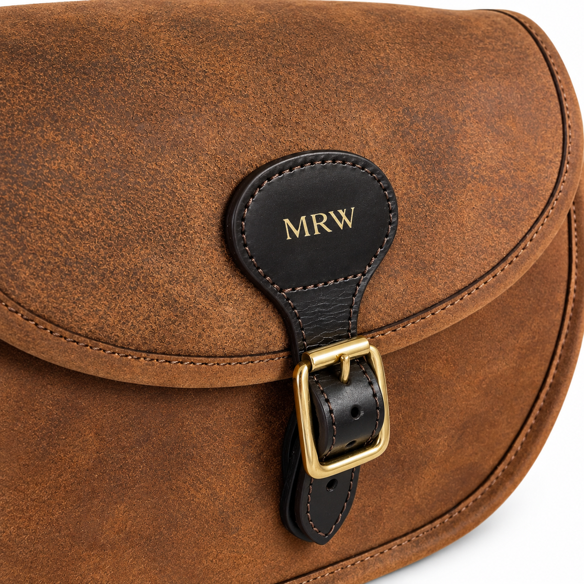 Close-up of a brown leather bag flap with a black leather strap closure and gold buckle. The black tab is personalised with the initials 'MRW' in gold.