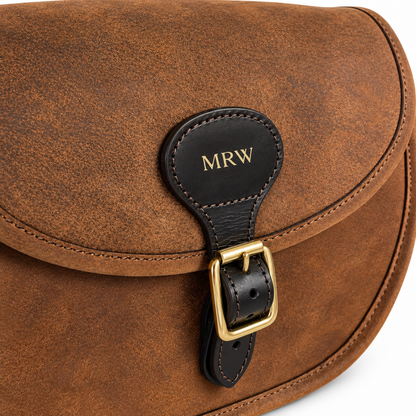 Close-up of a brown leather bag flap with a black leather strap closure and gold buckle. The black tab is personalised with the initials 'MRW' in gold.